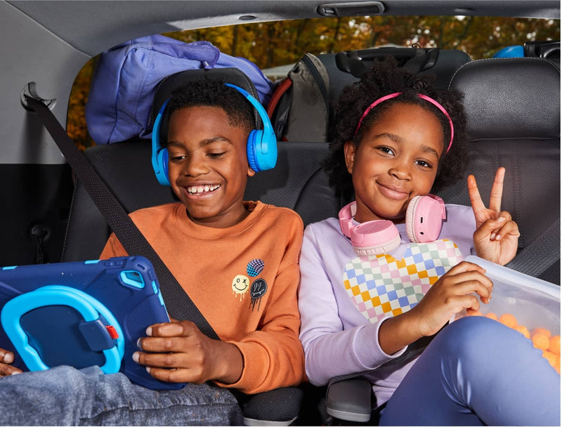 Two kids in a car: boy using a tablet, girl holding snacks, with luggage in the backseat.