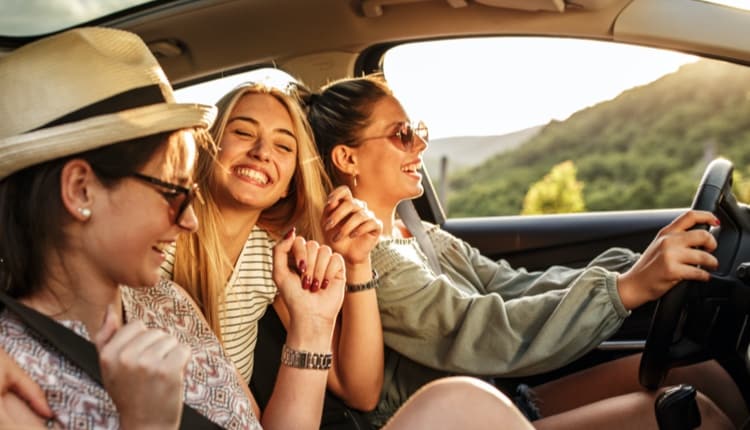 Three friends laughing in a car, enjoying a scenic drive in a sunny mountain setting.