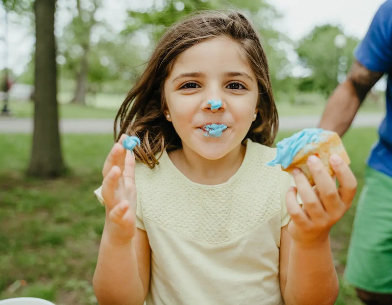 A child with frosting on their face smiles while holding a cupcake in a sunny park.