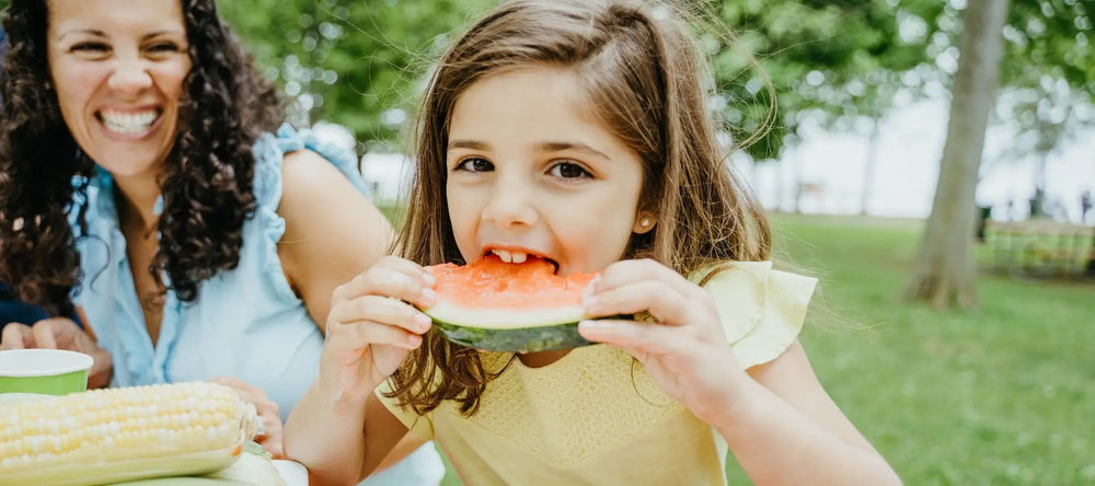 Smiling mother and daughter enjoying a picnic, with the girl eating a slice of watermelon outdoors.