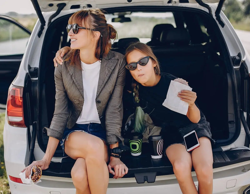 Mother and daughter sitting in the back of an SUV, enjoying snacks and drinks during a road trip.